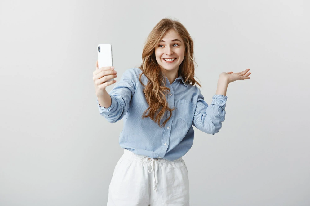 here-is-my-room-portrait-excited-happy-good-looking-woman-blue-blouse-showing-around-while-video-chatting-via-smartphone-smiling-broadly-directing-copy-space-gray-wall