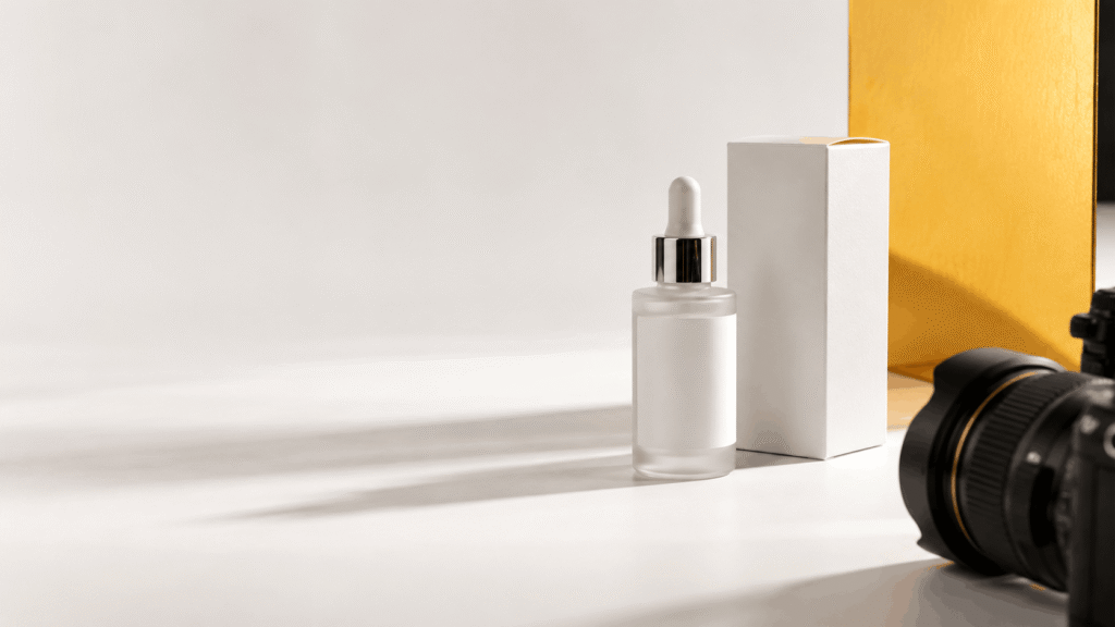A skincare bottle and packaging arranged beside a camera lens on a clean studio surface, ready for a product photo shoot.